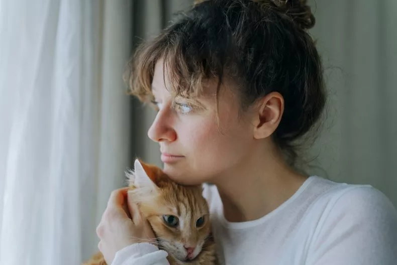 A woman looks out of the window holding her orange cat